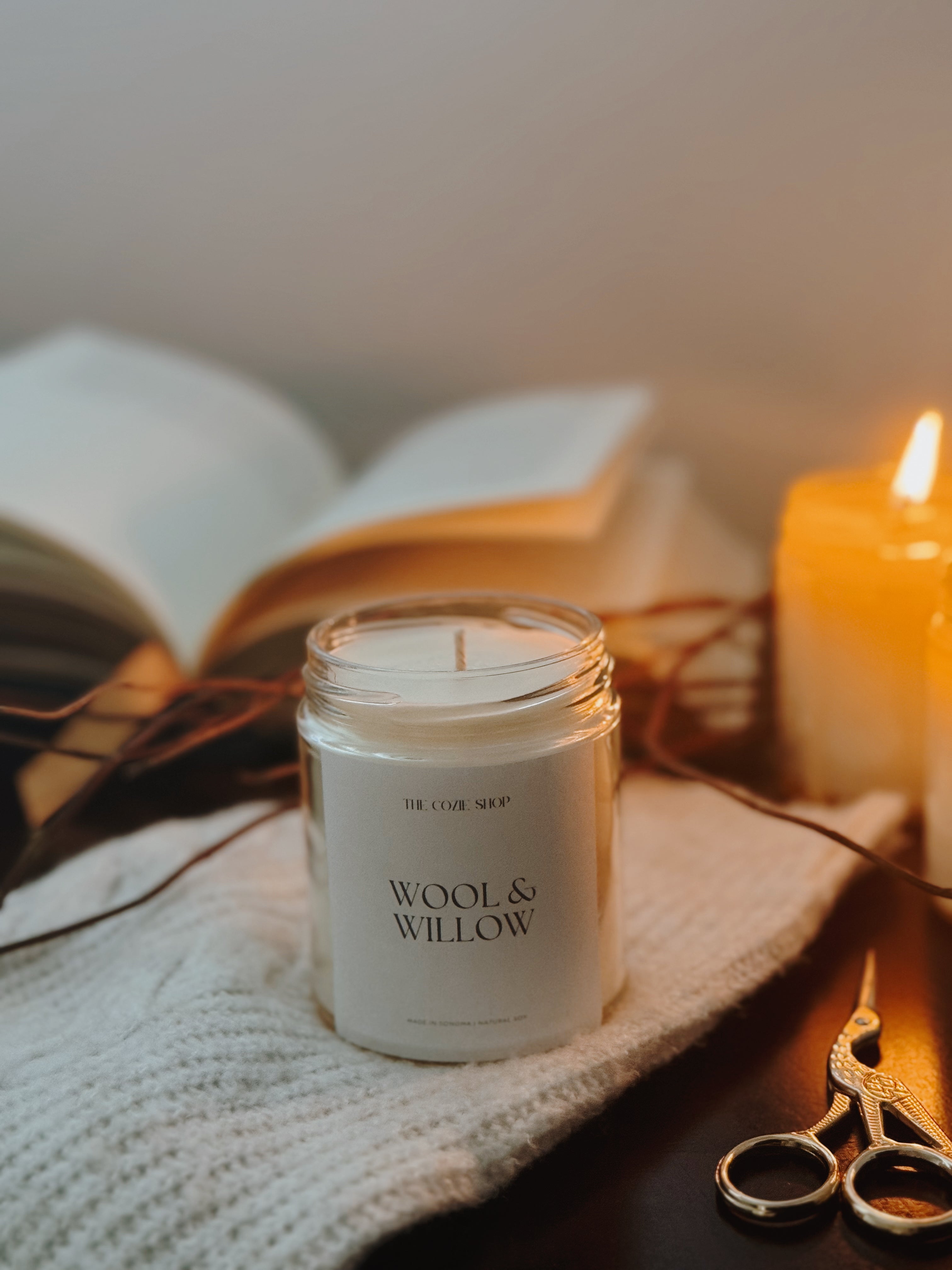 Wool & Willow candle on a surface with an open book and scissors in the background