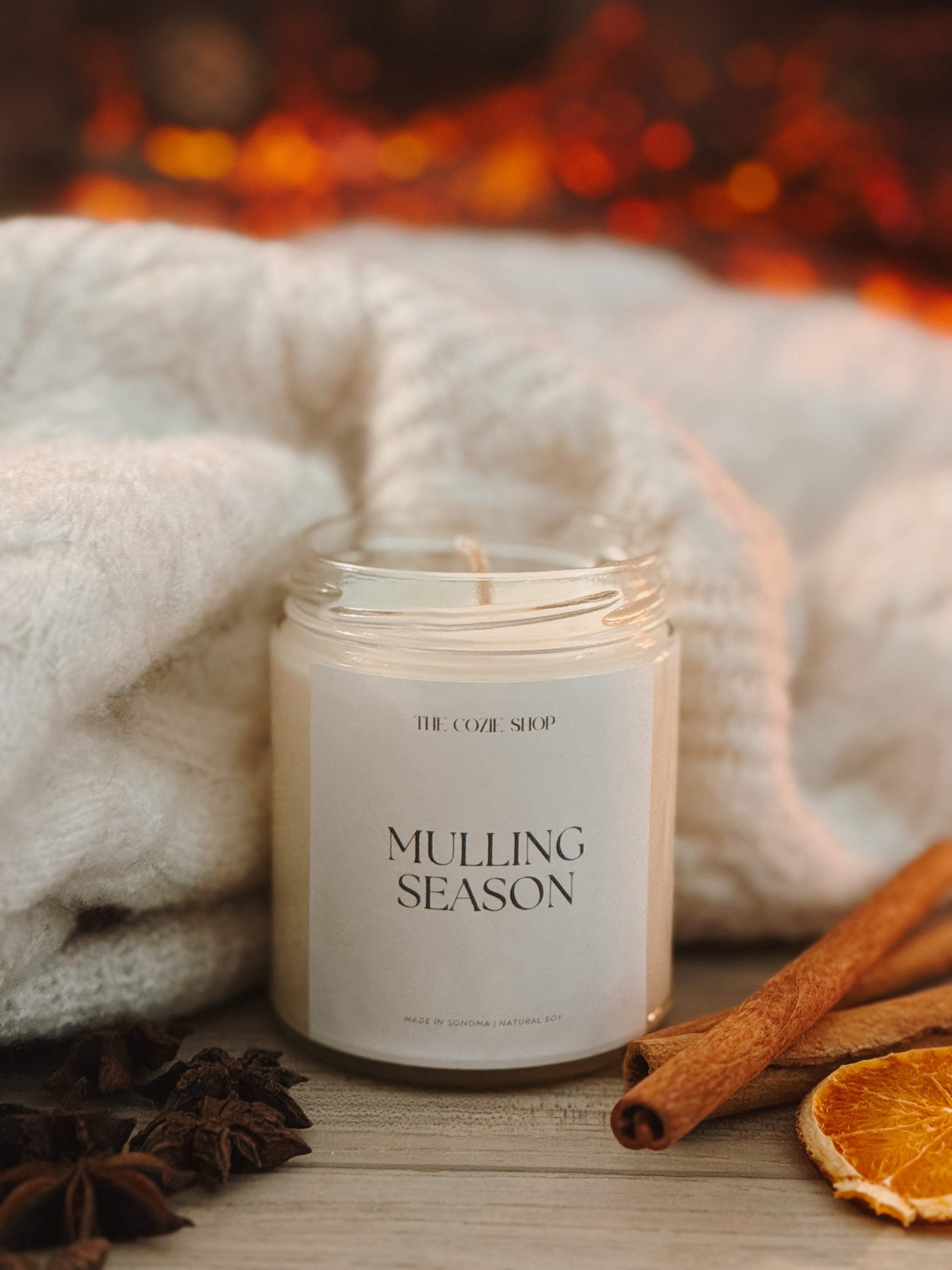 Candle labeled 'Mulling Season' with cinnamon sticks and star anise in front of a fireplace.