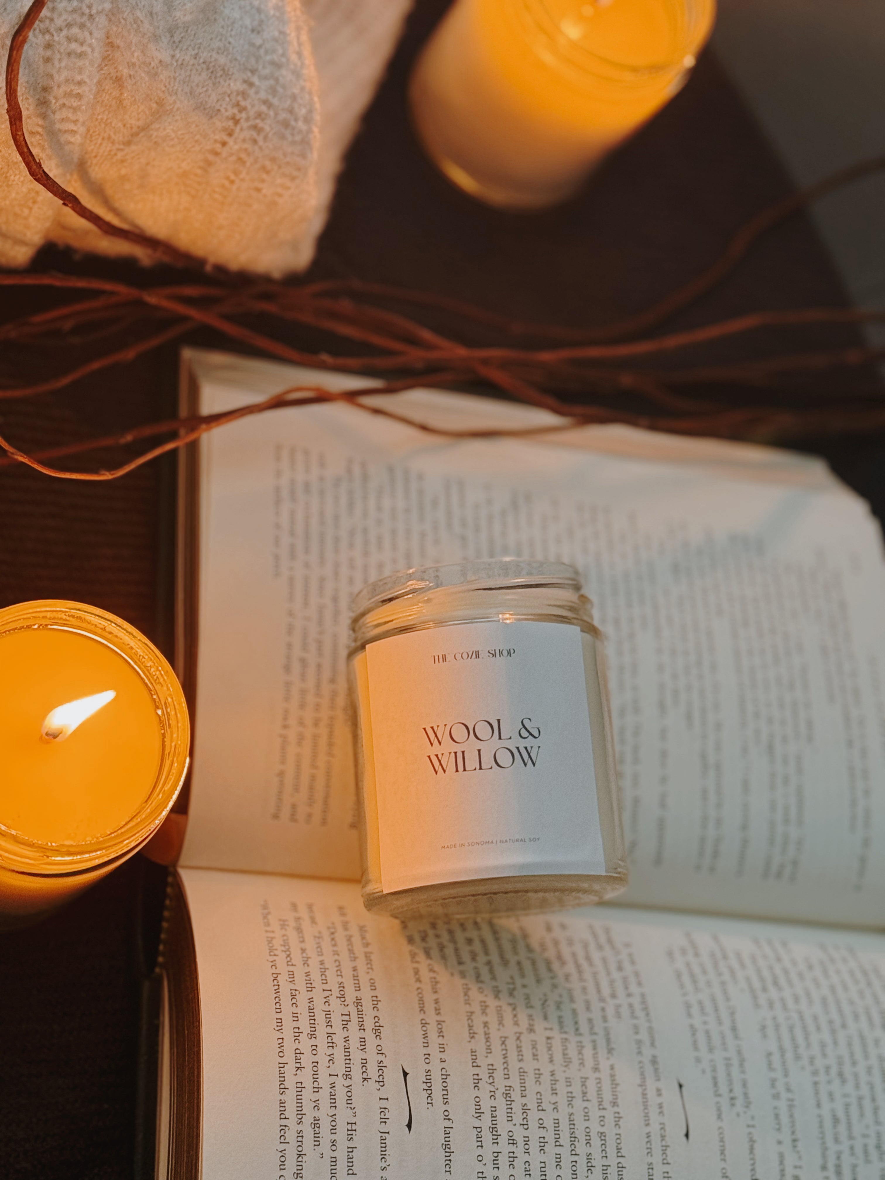 Wool & Willow candle on an open book with additional candles and string lights in the background.